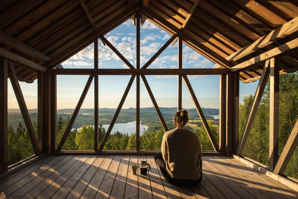A woman sits on the floor of an open loft in the morning light, with freshly brewn coffee at her side, overlooking the mountains and forest of Orbaden in Hälsingland by Martin Edström