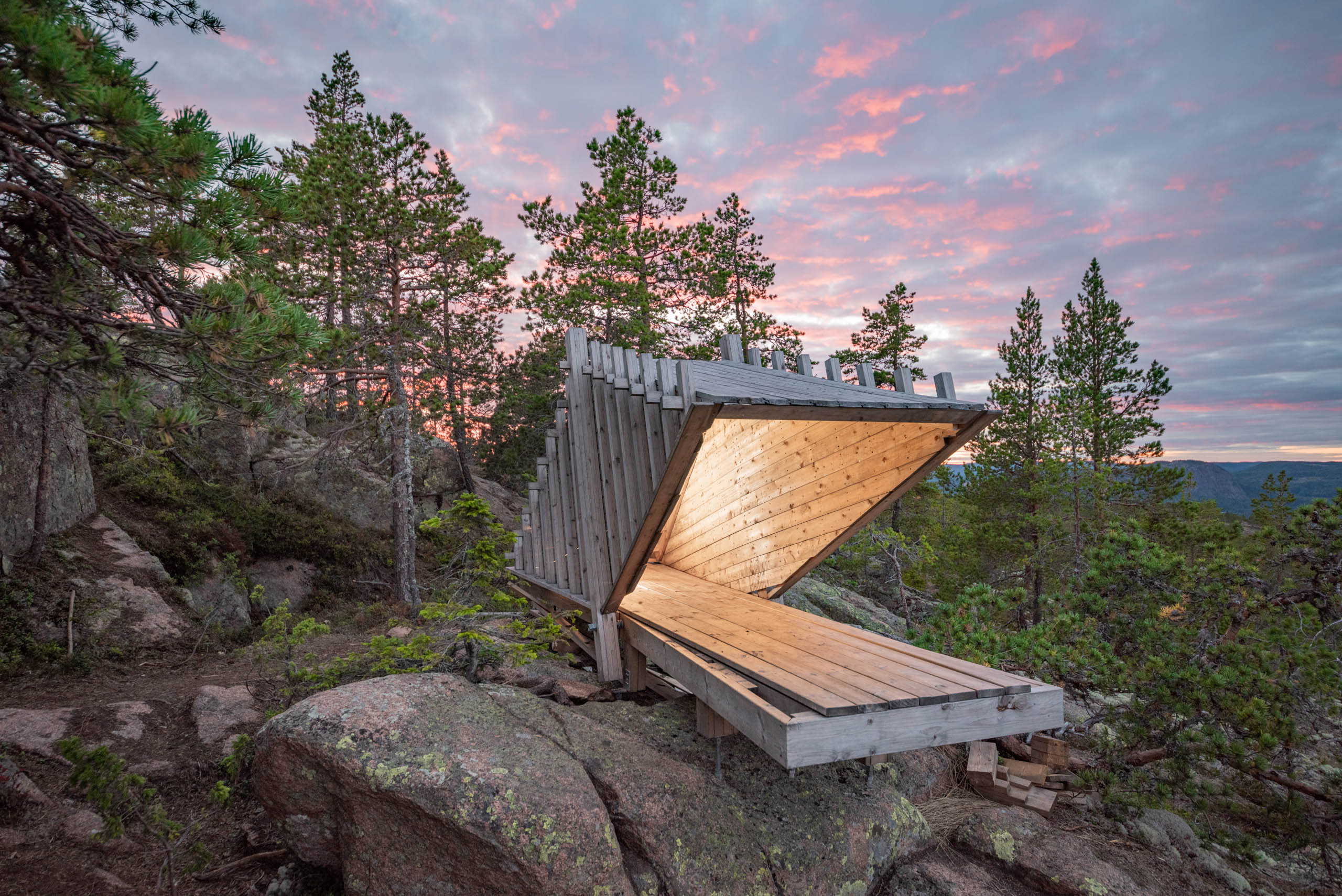 A hut or shack built with minimalistic architecture stands on top of a forested hill of the High Coast of Sweden, with a colorful twilight sky behind it by Martin Edström